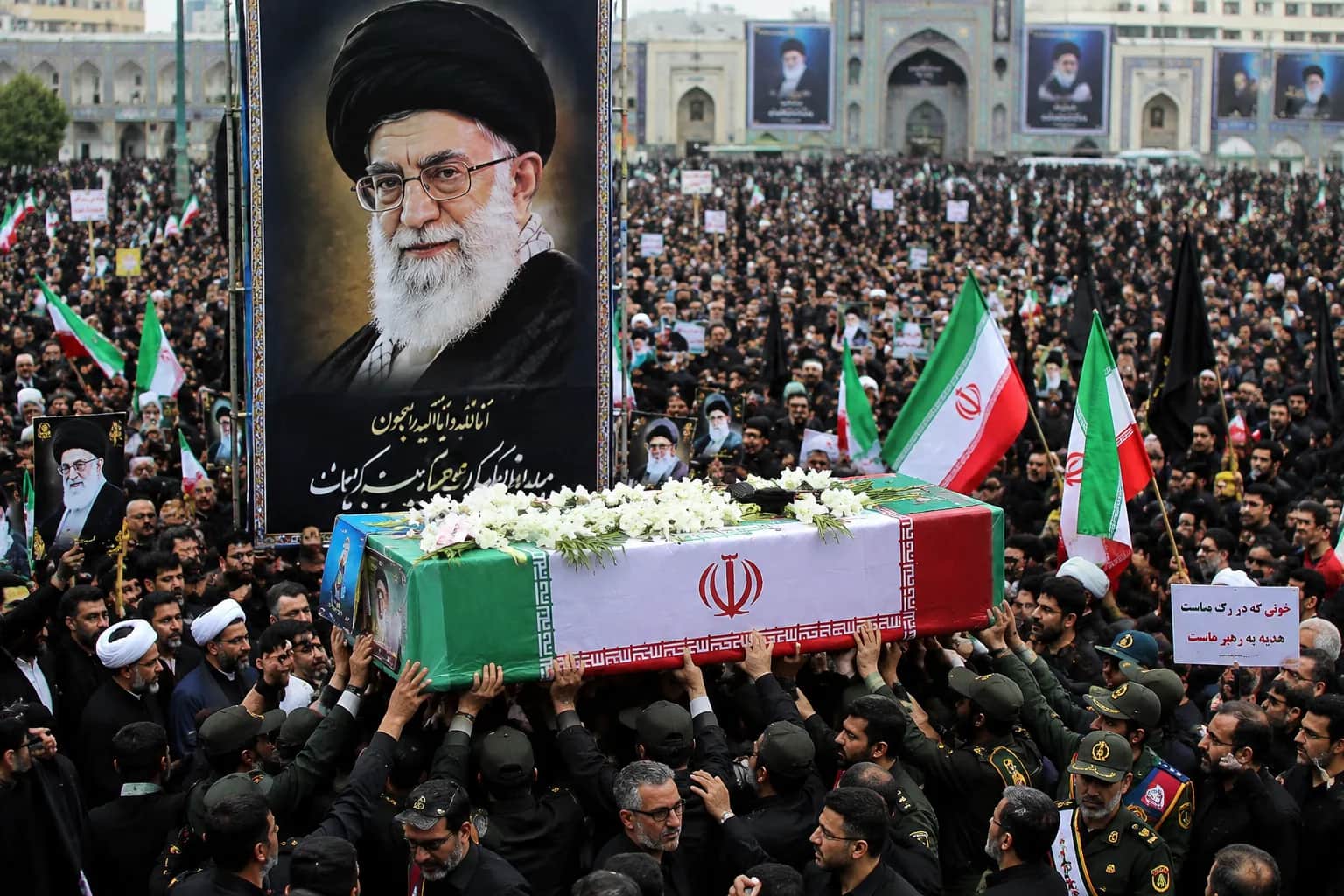 Thousands of mourners carrying Iranian flags surround the coffin of Ali Khamenei during a state funeral in Tehran, with security personnel, religious leaders, and black-clad crowds filling the ceremonial grounds.